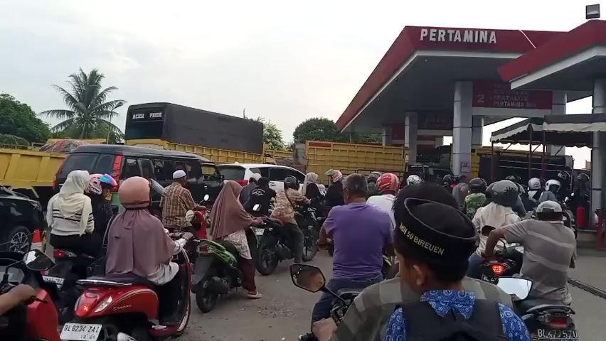 The video shows a traffic jam at a Pertamina gas station. Many motorcycles and a few cars are lined up, indicating long wait times. People are dressed in casual clothing, and some are wearing helmets. The environment appears to be outdoors with a cloudy sky. Trucks are visible in the background, suggesting a busy road or industrial area. The overall scene depicts a common traffic congestion scenario at a fuel station.