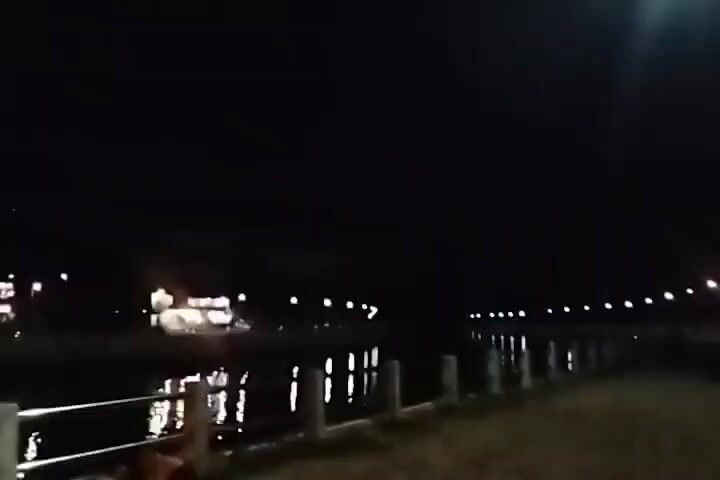 A nighttime video shows a waterfront path next to a dark body of water. Lights from buildings and streetlights are visible on the opposite bank, reflecting in the water. A metal railing runs along the edge of the path. The sky is black.