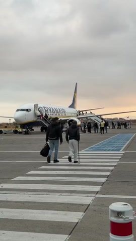 The video shows passengers disembarking from a Ryanair airplane on a tarmac. The plane is white with blue and yellow Ryanair branding. Passengers are walking down the stairs of the plane and across a crosswalk on the tarmac. Two men are walking away from the plane, one carrying a bag and the other using a walking stick. Other airport personnel are visible near the plane. The sky is overcast with a hint of sunset.