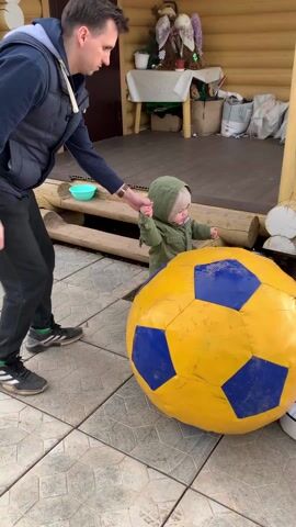 A man and a toddler are outside near a large yellow and blue soccer ball. The man, dressed in a dark vest over a dark blue hoodie and black pants, is holding the toddler's hand. The toddler, wearing a green hooded jacket, patterned pants, and a light beanie, is standing next to the soccer ball, touching it with their other hand. They are standing on a tiled patio next to a wooden building with stairs leading up to a porch. On the porch, there's a table covered with a white cloth and various items scattered around. The man appears to be guiding the toddler's interaction with the ball.