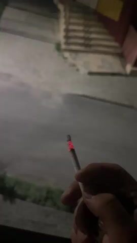The video begins with a close-up of a hand holding a lit cigarette. The background is out of focus, showing a dimly lit street with a building and stairs. The camera then pans out and moves, showing more of the street and the building. The building appears to be a multi-story structure with windows. There are streetlights visible, illuminating the scene. A car is parked on the side of the road in the distance. The video continues to show the street and building from different angles as the camera moves. The overall impression is a nighttime scene on a quiet street.