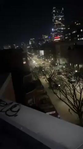The video depicts a nighttime urban scene from an elevated viewpoint, possibly a rooftop. Below, a street is visible, illuminated by streetlights, lined with bare trees and buildings of varying heights, including brick row houses and larger, modern structures in the distance. A tall skyscraper dominates the background, its lights twinkling against the dark sky. Sparse vehicle traffic can be observed on the street. The camera is initially stationary but then pans slightly to the right, revealing more of the cityscape. Graffiti is visible on the edge of the rooftop where the video is being filmed. The scene is generally dark with the city lights providing the primary source of illumination.