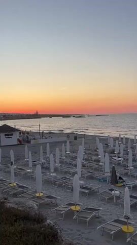 The video shows a beach with many empty lounge chairs and umbrellas. The sky is a gradient of orange and pink, indicating sunset or sunrise. The ocean is visible in the background, with gentle waves lapping at the shore. There is a small building on the left side of the frame that appears to be a beach hut or cabana. Some people are visible on the beach, but they are distant and their actions are not clear. The overall atmosphere is peaceful and serene.