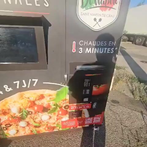 This is a close-up, slightly shaky video of a pizza vending machine. The machine is black with graphics of a pizza, the brand "Di Napoli," and text indicating pizzas are fresh, artisanal, and hot in 3 minutes, available 24/7. The machine has a screen, a payment terminal with a keypad, and a slot for dispensing. A person's shadow is visible on the machine and the ground, indicating their presence but they are not clearly visible. The vending machine is outdoors, with pavement and some vegetation visible in the background, along with a large truck. The lighting suggests it is daytime.