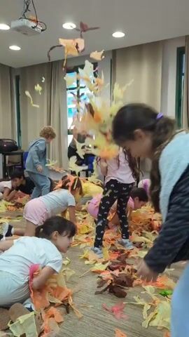 In a brightly lit, rectangular room with beige-toned walls and a beige floor, a group of about ten children are engaged in a playful activity, scattered across a floor covered in what appears to be orange and yellow shredded material. The children are dressed in a variety of casual clothing, including white t-shirts, pink pajamas, and gray jackets. They are kneeling, crawling, and walking through the shredded material, interacting with it by throwing it in the air and gathering it in their hands. An adult woman in black leggings and a green tank top stands near the back of the room, leaning over a table. There is a projector hanging from the ceiling and a speaker on the floor in the background. A window with green trim is visible in the background.