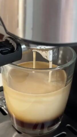This is a close-up, vertical video of an espresso shot being poured from an espresso machine into a clear glass mug. The espresso machine's portafilter is visible at the top of the frame, with two streams of espresso emerging from it and falling into the mug. The espresso fills the mug, creating a layered effect with a dark brown base and a lighter, creamy layer on top. The mug is filled to about two-thirds of its capacity. The background is blurred, suggesting a home or cafe environment. The video is characterized by its focus on the brewing process and the visual appeal of the freshly brewed espresso.