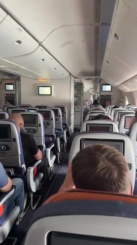 This video provides a first-person perspective from inside the passenger cabin of an airplane. The camera is positioned looking forward down the aisle. Rows of seats with individual entertainment screens are visible on both sides. Some seats are occupied by passengers, seen from behind. The lighting appears to be a mix of overhead cabin lights and natural light coming from the windows. Overhead compartments and emergency signage are also visible. The overall environment suggests a typical commercial flight.