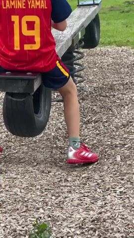 The video shows a young person from the back, wearing a red and yellow soccer jersey with the name LAMINE YAMAL and the number 19 on it, and blue shorts with yellow trim. The person is sitting on a seesaw made of wood and metal, with large springs underneath. The seesaw has a black tire as part of its structure. The person's legs are visible, wearing grey socks with a cartoon character design and red soccer cleats. The seesaw is located on a playground covered with wood chips. The background shows green grass and trees. The seesaw is moving up and down, indicated by the person's changing leg position and the movement of the seesaw.