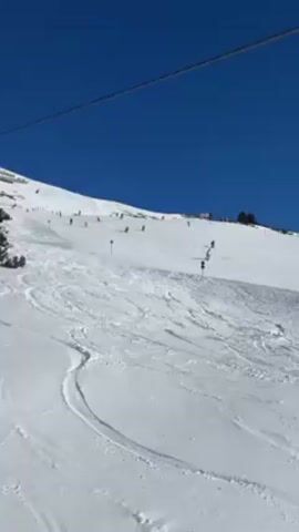 The video shows a snowy ski slope on a sunny day with a bright blue sky. Several skiers are visible descending the slope. A chairlift with empty chairs passes overhead. The snow is marked with ski tracks. Some trees are visible on the left side of the frame. There are blue trail markers on the slope. The video appears to be taken from another chairlift ascending the mountain.