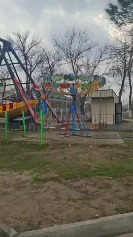The video shows an empty amusement park on an overcast day. The camera pans from right to left showing a colorful swing ride, then a Viking ship style ride, both unmanned, surrounded by trees without leaves. There’s a building in the background, a paved walkway in the foreground, and a red kiosk labeled “BOXER” on the left.