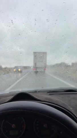 The video shows the view from inside a car driving on a highway in rainy weather. The windshield is covered in raindrops, and the windshield wiper is activated, clearing a portion of the view. In the distance, two trucks are visible on the wet road. The dashboard of the car, including the steering wheel and two gauges, is in the foreground. The sky is overcast and gray. The road appears to be a multi-lane highway with shoulders. The landscape on either side of the road is blurry and appears to be vegetation. The overall impression is one of driving in adverse weather conditions.