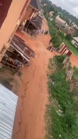 The video presents an aerial view of a bustling, unpaved street in Oko Town, Nigeria, during daylight hours. The road, characterized by its reddish-brown dirt and visible tire tracks, appears to be a central thoroughfare for local activity.

Several people, mostly men, are observed on the street. Some are gathered in small groups, seemingly engaged in conversation, while others are walking along the road. Two individuals are prominently seen standing next to motorcycles, with several other motorcycles and at least one bicycle parked nearby, suggesting these are common modes of transport in the area.

On the left side of the road, there's a simple structure with a corrugated iron roof, featuring a small, unreadable blue and white signboard. The road itself shows signs of natural wear, including what appears to be a dry, eroded channel running parallel to its left edge. The right side of the street is bordered by lush green vegetation, interspersed with visible litter such as plastic bags and bottles. The weather appears dry, and the overall scene depicts a typical, active street in a rural or semi-urban Nigerian setting.
