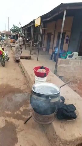 The video begins with a close-up of a motorcycle's handlebars and front end. The camera then pans right, revealing a dusty street scene with several motorcycles, cars, and a blue van. People are visible walking and standing, some near roadside stalls. A few motorcycles are parked along the street. The buildings lining the street appear to be made of brick and corrugated metal. The sky is overcast. As the video progresses, the camera continues to pan and move forward, showing more of the street and the activity on it. A person in a patterned dress stands near a stall on the right. A motorcycle with a red and yellow bag on the back is parked in the middle of the road. A small kiosk with a blue table is visible on the left, with a person in a yellow shirt standing behind it. Another motorcycle is parked in front of a building in the background. The video appears to be filmed from a moving vehicle or while walking.