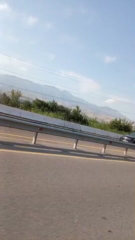 The video captures a view from a vehicle moving along a highway, likely in or near Gazalkent, Uzbekistan. The scene is dominated by a metal guardrail running alongside the road, with yellow lines marking the lanes. Beyond the guardrail, a landscape of rolling hills covered in lush green vegetation unfolds under a bright, partly cloudy sky, suggesting daytime and fair weather. In the distance, majestic mountains form a hazy backdrop.

Several power lines crisscross the sky, supported by tall, metal pylons, one of which is prominently visible on the right side of the frame. The presence of these pylon suggests proximity to an electricity infrastructure.

The primary activity is the movement of vehicles. A white minivan is seen driving in the opposite direction, its windows tinted. The driver is visible inside the minivan. The overall mood is neutral, depicting a typical journey on a highway with natural scenery. The footage appears to be taken during daylight hours, likely in the morning or afternoon, given the brightness and the clear sky. There is no visible text in the media.