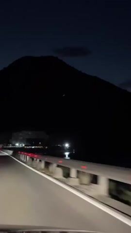 A car drives along a well-lit road at night. To the left, there's a body of water with lights reflecting off its surface. Behind the water, a dark mountain looms. Another car is driving ahead, its tail lights visible. A guardrail lines the side of the road.