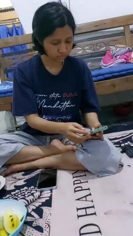 A young woman sits cross-legged on the floor, facing the camera and looking down at her phone, which she holds in her hands. She wears a dark blue t-shirt with white text that appears to say "New York State District Manhattan" and grey sweatpants. To her left, on the floor, is a white bowl containing yellow fruit. Also on the floor, directly in front of her, are two mobile phones, one black and one silver. The background shows a wooden bed frame with a blue mattress and some bedding. The lighting is dim and appears to be from an overhead source.