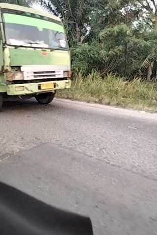 The video shows a view from the side of a vehicle, possibly a truck, with the road and foliage on the right side of the frame. The truck's tire is visible, and it appears to be in motion. The camera then pans to show two people on a motorcycle. The motorcycle is on its side as if it's losing control or performing a stunt. The rider and passenger are on the motorcycle, and it is moving on the road. The video continues to show the road and the trees and vegetation along the side of the road. The motorcycle appears to be attempting to correct itself, but it is still angled. The video briefly shows the front of the motorcycle before it moves out of frame. The environment appears to be rural, with lush greenery and a paved road.