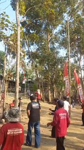 The video shows a group of people gathered outdoors, possibly for an event. They are positioned in a sandy area surrounded by trees. In the background, there is a volleyball net stretched between two poles. Several people are visible on both sides of the net, and some are in motion, suggesting they are participating in or observing an activity. Red and white flags, some with text, are prominently displayed on poles, adding a festive or celebratory atmosphere. The lighting indicates it is daytime, likely with clear skies. Some individuals are wearing red shirts, while others are in casual attire. The overall scene appears to be a community gathering or a sporting event.