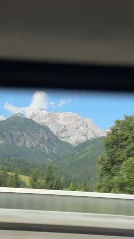 A car is driving through a mountainous landscape with lush green trees in the foreground and a large, rocky mountain range in the background under a clear blue sky with a few white clouds. The camera is looking out of the car window, and the trees are blurred due to the motion. There are no people or animals visible in the frame. The time stamp indicates the video is a short clip.
