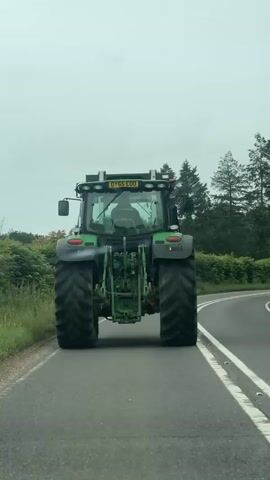 A green John Deere tractor is driving away from the camera on a paved road. The tractor has large rear tires and appears to be on a rural road with trees and bushes on either side. The sky is overcast. The tractor has its hazard lights on. The license plate is visible and reads DY65 EDO. The tractor appears to be moving at a moderate speed.