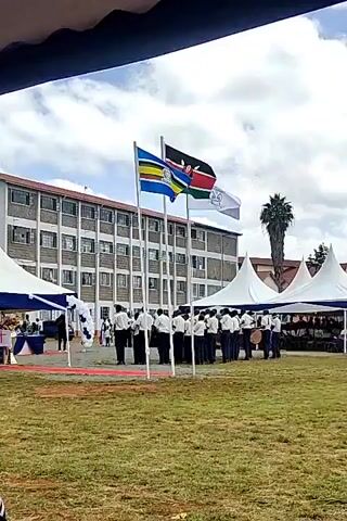 The video shows an outdoor event on a grassy field. In the foreground is a group of people in white shirts and black pants, appearing to be a band, standing in front of three flagpoles. The flags from left to right are a blue, yellow, red, black and white flag, the Kenyan flag, and a white flag. Behind the group is a multi-story brick building and a row of white tents. A palm tree is visible towards the right side of the frame. The sky is partly cloudy.