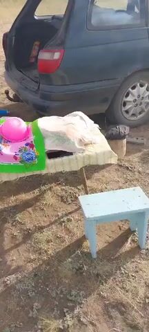 The video shows an outdoor scene with a car trunk open in the background. On a table covered with a colorful cloth, a pink bowl and a white towel are visible. Nearby, there's a blue stool and a water bottle. To the right, a silver samovar is standing on the ground next to a blue plastic bag. A large white bag filled with dark objects is in the foreground. The ground is dry and dusty with some sparse vegetation.