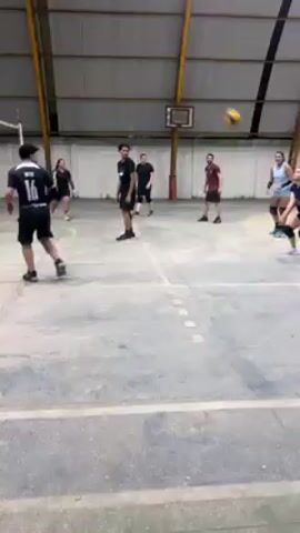 Several people play volleyball in an indoor gymnasium. They're split into two teams and are practicing serves, bumps, and hits.  The gym has a high arched roof, basketball hoops on the walls, and is lit by overhead lights. The floor is grey concrete marked with white lines. One person wears the number 16 on their back.