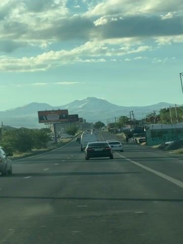 The video shows a view from a car driving on a highway. The highway has multiple lanes and is flanked by vegetation and some buildings on the right side. There are several cars on the road, including a black sedan in the foreground. In the distance, a snow-capped mountain is visible under a cloudy sky. Billboards are also present along the side of the road. The overall scene suggests a daytime drive on a suburban or rural highway.