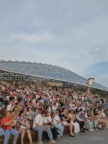 The video shows an outdoor jazz festival. A large crowd of people is seated on tiered bleachers in the foreground, facing a stage in the background. On the stage, a band is performing, with several musicians playing instruments. The stage is covered by a large, arched structure with lighting equipment. The sky is visible above, with some clouds. The overall impression is of a lively outdoor event.
