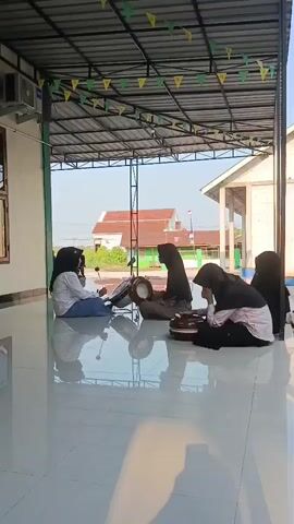 Five individuals, all wearing hijabs, are sitting on a shiny tiled floor under a covered outdoor area. The individuals appear to be playing musical instruments. The person on the far left is holding a stick and appears to be striking a drum. To their right, another person is holding and playing a drum. The three individuals further to the right are also holding and playing round, flat percussion instruments, possibly tambourines or frame drums. The environment appears to be a school or community building, with a building visible in the background. Festive triangular flags are strung across the ceiling of the covered area. The floor is highly reflective, showing the reflections of the people and the overhead structure. The lighting suggests it is daytime, with sunlight casting shadows.