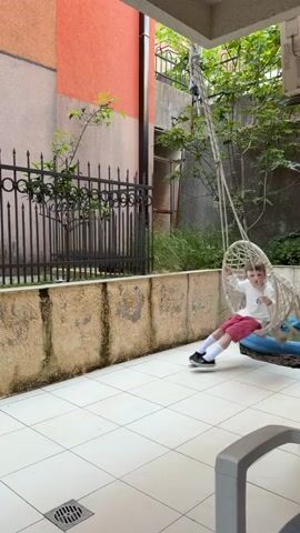 A young child swings on a hanging hammock swing in a backyard setting. The child is wearing a white top, pink shorts, white socks, and black shoes. The swing hangs under a covered patio area with white tiled floors.  Behind the swing, there is a small, blue inflatable pool along a low, weathered wall topped with an ornate black iron fence.  Behind the fence, greenery grows.  The walls of the surrounding houses are painted in shades of peach and beige. The camera remains stationary throughout the video.
