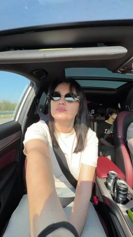 A woman with dark hair and sunglasses is in the driver's seat of a car. She is wearing a white t-shirt and a seatbelt. She looks at the camera and touches her hair. There are two cans of drink in the center console of the car. In the back seat, another person is visible. The car is on a road with trees and blue sky visible through the windows and sunroof.