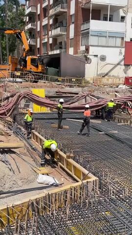 A construction site scene unfolds with multiple workers engaged in various tasks. In the foreground, a worker in a bright green shirt and white hard hat is bent over, working with rebar and formwork. To the left, another worker in a similar outfit is standing and appears to be supervising or performing a task. In the mid-ground, three workers are visible, one in a yellow vest and white hard hat, another in an orange shirt and white hard hat, and a third in a green shirt and white hard hat. They are standing amidst rebar grids and large red hoses. In the background, an orange crane is positioned, and a multi-story building with balconies is visible. The environment is an active construction zone with dirt, rebar, wooden forms, and various construction materials and equipment. The overall impression is one of active construction work in an urban setting.