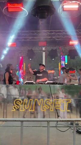 A group of people are on a stage in front of a DJ booth. The DJ is a man wearing a black t-shirt and a baseball cap, and he is playing music. There are several other people on the stage with him, including women wearing tank tops and shorts. In the foreground, there is a neon sign that says SUNSET. The stage is lit with red and white lights, and there is smoke in the air. The event appears to be an outdoor music festival or party.