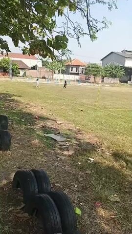 The video shows an outdoor field with a grassy surface. Several tires are stacked on the left side of the frame, near some trees and dry foliage. In the background, there are buildings, some with orange roofs, and a chain-link fence. Two figures are visible on the field; one appears to be running, and the other is walking. A yellow pole is planted in the grass. The sky is bright and clear, suggesting a sunny day. The overall scene depicts an open area that could be a park or a sports field.