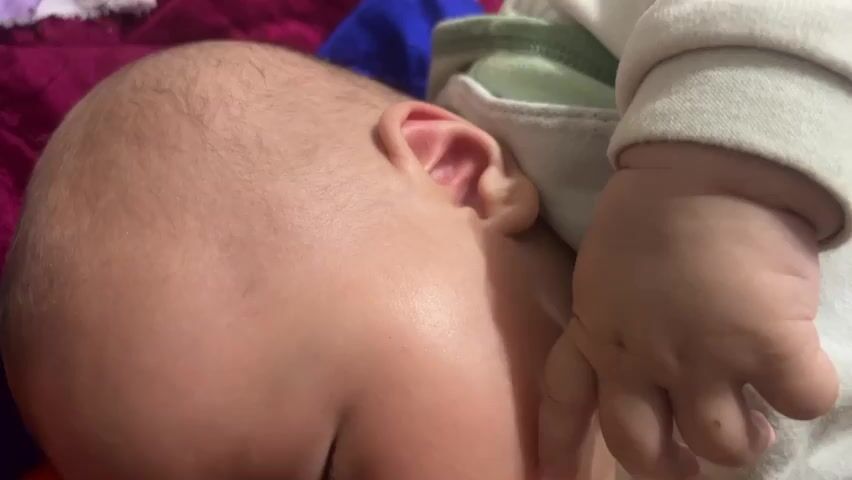 A close-up shot of a baby sleeping on its side. The baby's head is turned to the left, with its eyes closed and its mouth slightly open. A small hand is resting near the baby's face. The baby is wearing a light-colored onesie. The background is blurred but appears to be a bed with blankets. The lighting is soft and natural.