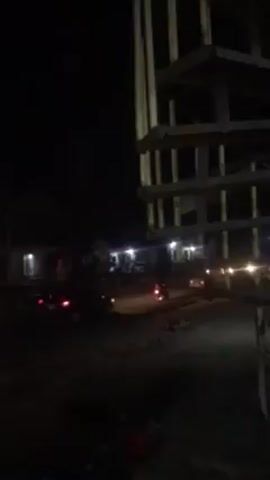 The video depicts a street scene at night. Several vehicles, including auto rickshaws and possibly cars, are visible on the road, with their headlights illuminating the scene. There are a few buildings on the side of the road, with lights emanating from their windows. A tall, skeletal structure stands prominently in the foreground, partially obscuring the view of the street. The lighting is dim, with the primary sources of illumination being the vehicle headlights and the lights from the buildings. A small child is seen on the side of the road.