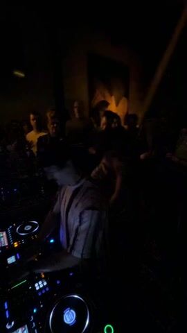 The video shows a DJ in a striped shirt playing music for a crowd at a nightclub. He is using a modern DJ console with multiple knobs, dials, and screens that emit green and blue light. Several people are standing behind a bar, and others stand further back, some appearing to dance. The lighting in the club is generally dim. Someone is standing close to the DJ, with their arm raised in the air, as if dancing or enjoying the music. The rest of the crowd is visible as silhouettes in the dark.