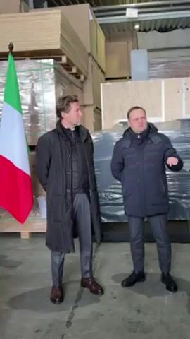 Two men stand in a warehouse near pallets of wooden boards. To their left is the flag of the European Union, and the flag of Italy. The man on the left is wearing a long black coat, and gray pants. The man on the right is wearing a puffy, dark blue coat and gray pants. The man on the right is gesturing with his right hand as he is speaking.