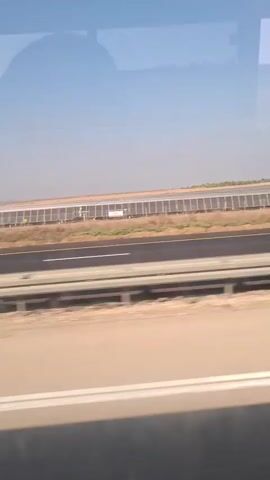 The video shows a view from a moving vehicle. A highway is visible in the foreground with a truck and a car driving on it. In the background, there is a large solar farm with rows of solar panels. The sky is clear and blue.