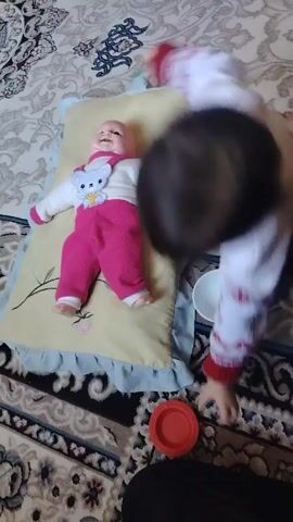 The video shows a child playing with a doll lying on a pillow. The doll is wearing a white sweater with a bear design, a pink scarf, and pink overalls. The pillow is beige with a blue ruffle. The child, wearing a white sweater with a red pattern and dark pants, is kneeling next to the doll and using a green stick to poke at the doll's face. Next to the child are a small white bowl and a red plate. The scene is set on a patterned rug with beige, gray, and black designs.