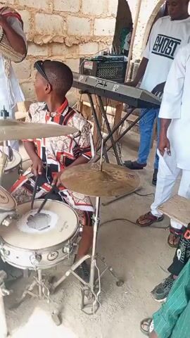 In an outdoor setting under a makeshift roof, a young boy plays a drum set. He is wearing a colorful, patterned shirt and shorts, with a headband. Another man stands nearby, playing a hand drum and singing into a microphone. He is dressed in a white outfit.  A keyboard and electronic equipment are visible behind them.  Other children are present, watching the musicians. The environment appears to be a casual, possibly residential area.