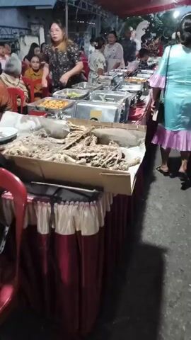 The video shows an outdoor buffet at an event. In the foreground, a long table is covered with a red and white tablecloth, laden with chafing dishes of food. Some dishes appear partially eaten or nearly empty, with visible remnants of food. There are also some raw ingredients or prepared dishes on a cardboard surface in front of the table. People are gathered around the buffet, reaching for food and interacting. The background shows more people, some seated at tables, and a canopy or tent overhead. The lighting suggests it is evening or dusk.