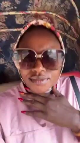 A woman wearing sunglasses and a headscarf is the subject of this close-up video. She is wearing a pink long-sleeved shirt, and has a black purse strap over her shoulder. She is inside a vehicle with a patterned, floral roof. The woman looks at the camera with a slight smile. Her fingernails are painted red. The camera is slightly shaky, suggesting movement.