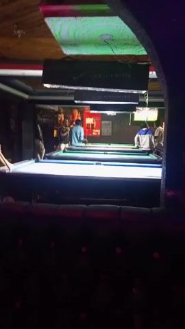 A pool hall is the setting for this video. The scene is dimly lit with colored lights enhancing the mood. The camera is positioned near a padded booth, giving a partial view of the playing area. Several individuals are visible, engaging in games on multiple pool tables. A man in a dark hoodie stands on the left, holding a pool cue and taking aim. Further into the room, another person is seen lining up a shot, wearing a light blue shirt. A third person is on a pool table near the back right. Overhead, illuminated signs display text, and the ceiling has a decorative wood paneling design. The atmosphere suggests a relaxed, social environment centered around the game of pool.