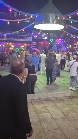 The video shows a crowded outdoor space at night, decorated with colorful string lights. In the foreground, a man in a black suit is walking away from the camera. In the midground, a group of people are gathered, some dancing. In the background, there are more people and some structures with lights, possibly shops or stalls. The ground is paved with square tiles.
