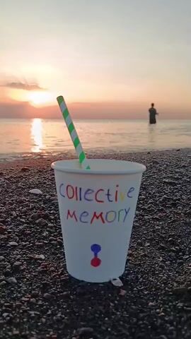 A close-up shot focuses on a white paper cup on a pebbly beach. The cup has the words "COLLECTIVE MEMORY" written on it in colorful letters, with a simple drawing of a blue and red icon below the text. A green and white striped straw is inserted into the cup. In the background, a calm ocean stretches towards the horizon, where the sun is setting, casting a warm orange glow. A lone figure stands in the shallow water, appearing to be fishing with a rod and reel. The overall scene evokes a peaceful and contemplative atmosphere.