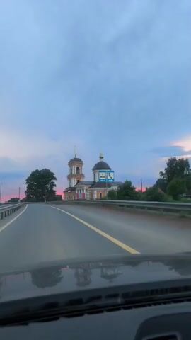 A dash cam records a forward view of a road passing by a yellow church with two domes and a bell tower. The church is situated on the right side of the road behind a metal guardrail. A large green tree is visible to the left of the church, and further to the right, a pinkish-red structure is partially visible. The sky is a mix of blue and pinkish hues, suggesting it is either sunrise or sunset. Road signs are present next to the church, one of which displays a phone icon. The road itself is paved and has a yellow line marking the lane. The bottom of the frame shows the car's dashboard.