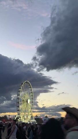 The video shows a fairground Ferris wheel at dusk. The sky is partially cloudy with colors of blue, pink, orange, and grey. The Ferris wheel is illuminated with white lights and is positioned in the background, behind a crowd of people. Individual people are visible in the foreground, including a man wearing a cowboy hat. The camera is moving slightly, causing some blur in the image.