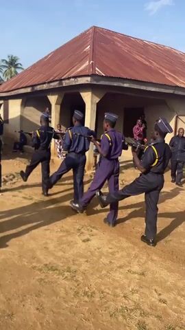 A group of individuals dressed in navy blue uniforms with blue caps are performing a synchronized marching drill outdoors in front of a building with a rusty corrugated metal roof. Some individuals are carrying what appear to be rifles. The ground is dirt and appears to be uneven. The sky is blue with some clouds. There are trees visible in the background. One individual in the foreground is facing away from the camera and appears to be rubbing their face. The group is moving in unison.
